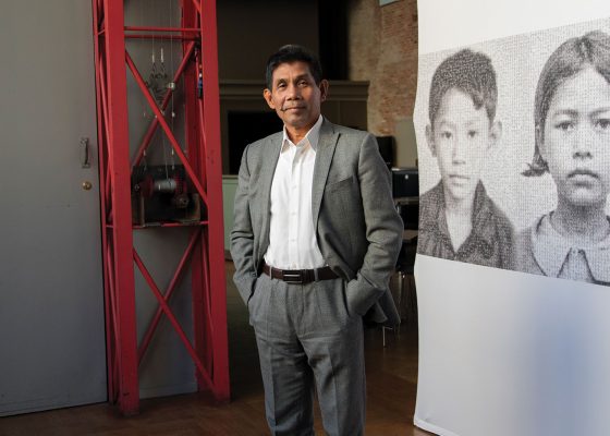 Him Sophy stands beside photo collage images of Khmer Rouge victims at the Brooklyn Academy of Music in Brooklyn, New York, 2017.
