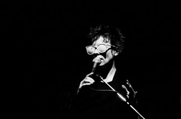 Laurie Anderson wearing glasses, singing into microphone
